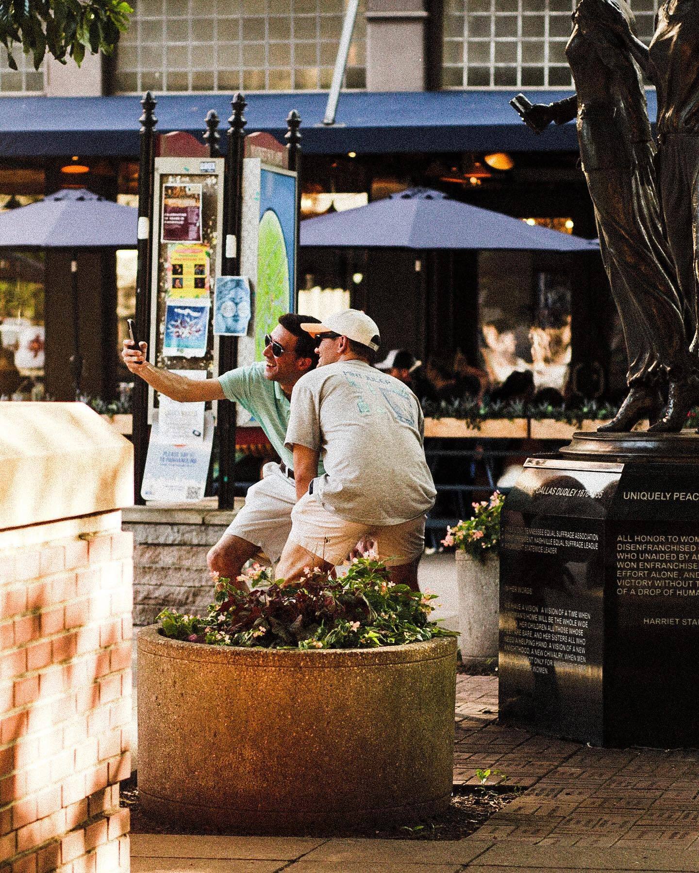 People at a café table under umbrellas, beside a bronze statue
